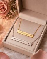 Two gold and silver necklaces with engraved coordinates in a jewelry box, surrounded by pink roses and petals.